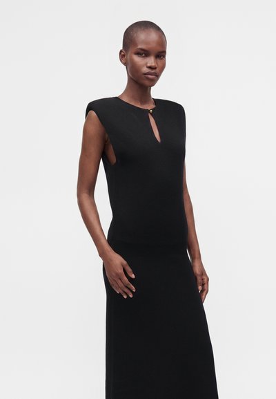 Femme mince aux cheveux courts portant une robe noire sans manches avec une petite ouverture devant et un bouton doré, posant devant un fond uni.