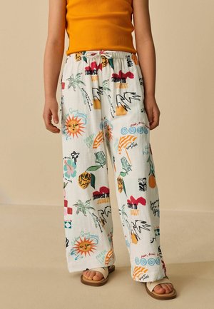 Kind trägt weiße, lockere Hose mit bunten sommerlichen Motiven und ein oranges ärmelloses Oberteil, steht mit Sandalen auf einem beigen Boden.