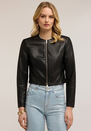 Frau trägt eine schwarze, kurze Lederjacke mit Frontreißverschluss, hellblaue hoch taillierte Jeans, ein goldenes Armband und goldene Ohrringe.