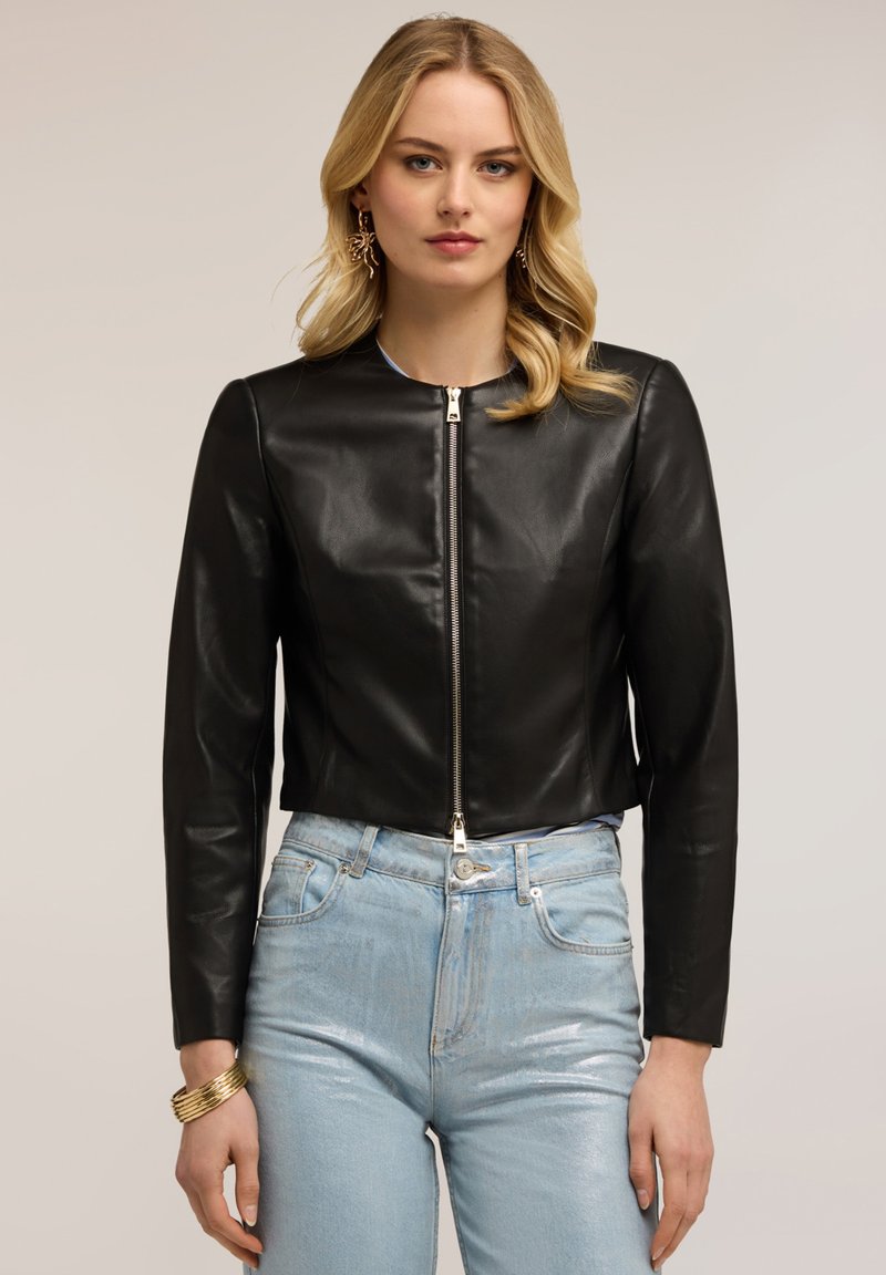 Donna che indossa una giacca corta in pelle nera con cerniera frontale, jeans a vita alta azzurri, braccialetto d'oro e orecchini d'oro.