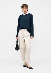 Personne debout portant un pull bleu marine, un pantalon large blanc, des ballerines noires pointues, et tenant un sac à main noir sur un fond uni.