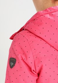 Giacca a vento rosa con piccole pois neri, dotata di un patch in tessuto sulla manica e dettagli di cucitura eleganti sul colletto.
