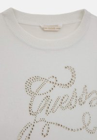 White sweater featuring a ribbed neckline and embellished with gold studs forming the word "Guess" in a flowing script design.
