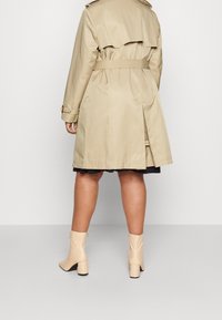 Person som bär en beige trenchcoat med skärp, svart kjol och beiga ankellånga stövlar, står på ett slätt ljusgrått golv mot en grå vägg.