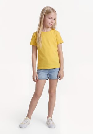 Mladi djevojka stoji, nosi žutu majicu kratkih rukava, svijetlo plave traper kratke hlače i bijele tenisice, gleda dolje i u stranu.