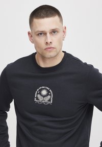 Fekete pulóver kerek nyakkal, fehér hegyek és nap grafikával. Puha anyagból készült, laza szabással.