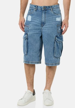 Mann trägt kniehohe blaue Denim-Cargoshorts mit mehreren Taschen und weißen Sneakers, steht vor einem einfarbigen Hintergrund.
