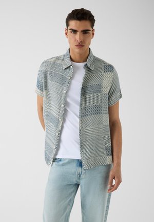 Jeune homme portant une chemise à manches courtes à motifs boutonnée par-dessus un t-shirt blanc avec un jean bleu clair sur un fond uni.
