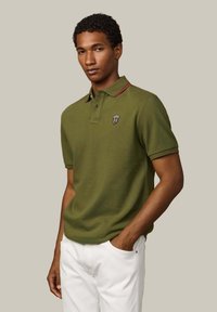 Olivovo zelené polo tričko s červeným pruhovaným detailom na golieri a logom na hrudi, vyrobené z bavlny. Model má na sebe biele nohavice, ruky v vreckách.