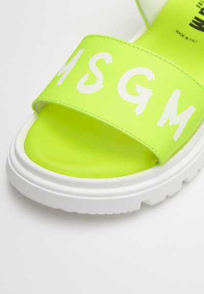 Νεόν πράσινη μπαντάνα με φαρδύ λουρί που διαθέτει λευκά χειρόγραφα γράμματα. Παχύ λευκό πέλμα με υφή για πρόσφυση.