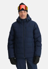 Navyblaue Steppjacke mit Kapuze, ausgestattet mit einem Reißverschluss vorne und seitlichen Taschen. Getragen mit Handschuhen und Skibrille wirkt der Stoff isoliert.
