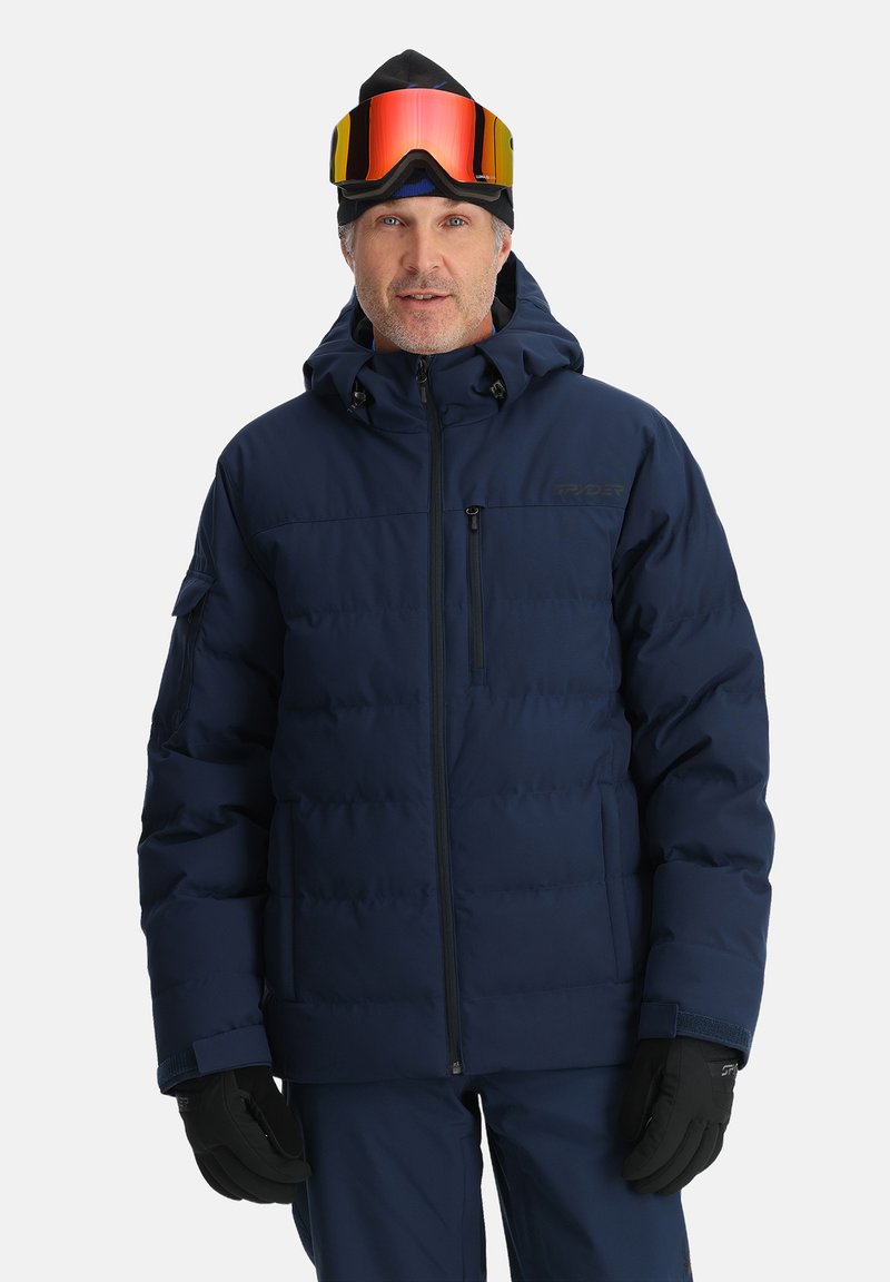 Navyblaue Steppjacke mit Kapuze, ausgestattet mit einem Reißverschluss vorne und seitlichen Taschen. Getragen mit Handschuhen und Skibrille wirkt der Stoff isoliert.