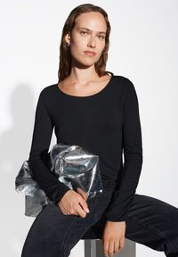 Schwarzes Langarmoberteil mit rundem Ausschnitt, kombiniert mit einer zerknitterten metallicsilbernen Tasche. Model sitzt und trägt dunkle Jeans. Schlichtes, elegantes Design.