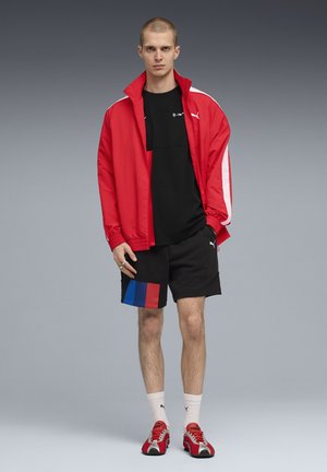 Joven de pie vestido con una chaqueta roja, pantalones cortos negros con parches rojos y azules, camiseta negra, calcetines blancos y zapatillas rojas sobre un fondo gris.