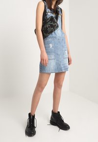 Denimoverallklänning i ljusblått med framskjutna fickor och fransade detaljer, kombinerad med en svart camouflagedesignväska och svarta snörboots.