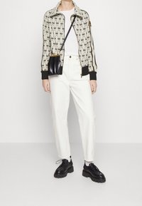 Chaqueta beige con cierre y un patrón negro, camiseta blanca, pantalones blancos y zapatos negros de suela gruesa. Bolso cruzado negro con detalles en dorado.