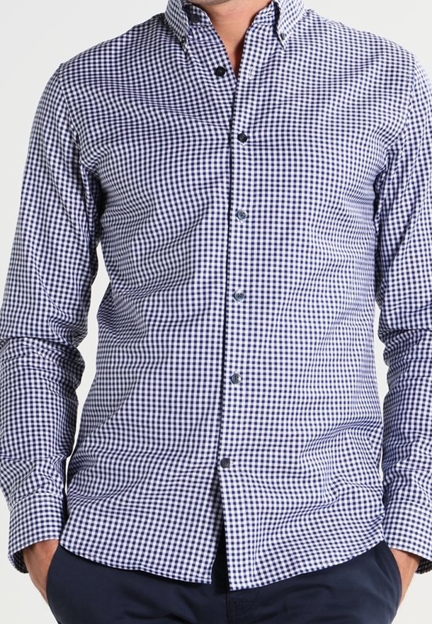 Man wearing a fitted navy and white checkered button-up shirt with hands in pockets, shown from neck to hips.