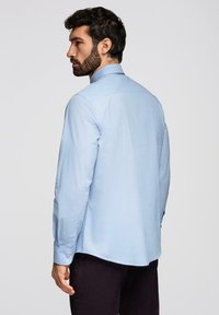 Homme aux cheveux foncés et à la barbe, portant une chemise bleue claire à manches longues boutonnée et un pantalon sombre, de profil sur un fond uni.