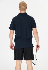 Mörkblå pikétröja med korta ärmar, av bomullsmaterial, och sidslitsar, ihop med svarta träningsshorts och ett badmintonracket.