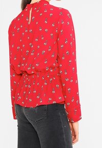 Röd blommig blus med insydd midja och en nyckelhålsknäppning på baksidan. Tyget är lätt och har små vita och gröna blommönster.