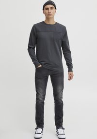 Maglietta grigia a maniche lunghe con un pannello superiore texturizzato, tasca frontale e scollatura rotonda, abbinata a jeans scuri e sneakers bianche.