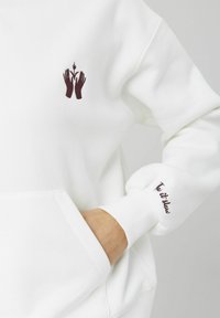 Sweat blanc avec des mains brodées en marron tenant une fleur sur la poitrine et le texte "Too it slow" sur la manchette, main dans la poche.