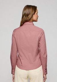 Camicia a maniche lunghe in cotone rosa polveroso, caratterizzata da un colletto a punta, polsini con bottoni e una silhouette aderente. La vista posteriore rivela un orlo curvo.