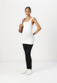 Witte mouwloze top met een gebogen ontwerp, zwarte legging en witte sneakers. Model houdt een bruine schoudertas vast en staat op een lichte achtergrond.