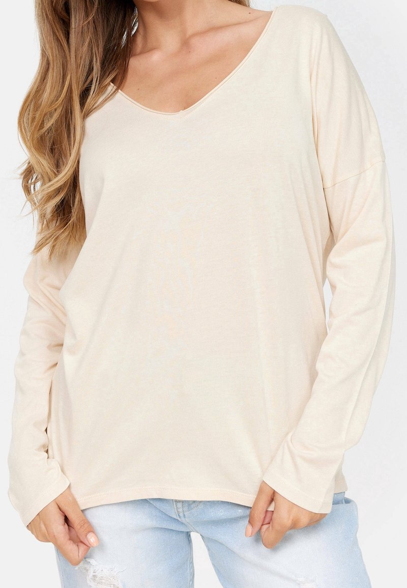 Beige långärmad topp med djup V-ringning, gjord av mjukt tyg. Har en avslappnad passform och är kombinerad med ljusblå slitna jeans.