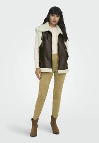 Bruine leren mouwloze vest met een licht nepbont kraag, gedragen over een lichtroze trui en beige suède broek, gecombineerd met bruine enkellaarsjes.