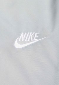 �Λευκό κεντημένο λογότυπο "Nike" σε φόντο απαλής, ανοιχτογκρι υφής. Το λογότυπο παρουσιάζει καμπυλωτό σχέδιο με έντονο σχήμα swoosh.