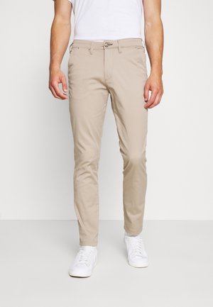 Mand iført beige slim-fit bukser, hvide sneakers og hvid t-shirt, stående mod en ensfarvet hvid baggrund.