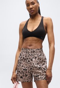 Γυμνασμένη γυναίκα με μαύρο αθλητικό σουτιέν και μπεζ σορτς με λεοπάρ σχέδιο στέκεται κρατώντας μια ροζ λαστιχένια ταινία αντίστασης μπροστά σε απλό φόντο.