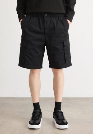Black cargo shorts made of cotton, featuring an elastic waistband, side pockets, and a relaxed fit. Worn with black socks and sneakers.