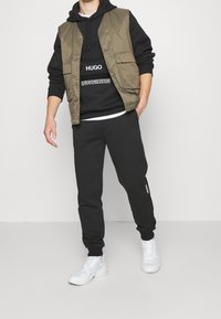Zwarte hoodie met witte tekst, gedragen onder een olijfgroene vest; gecombineerd met zwarte trainingsbroek en witte sneakers. Minimalistisch ontwerp.