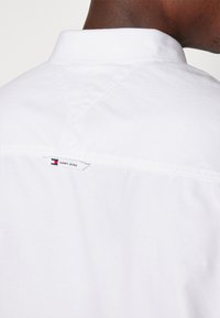 Maglietta bianca in cotone con colletto a punta, caratterizzata da una piccola etichetta Tommy Jeans sul retro. Linee pulite e una superficie liscia.