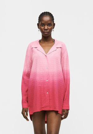 Kobieta z warkoczami, ubrana w różową koszulę na guziki z ombre i subtelnym wzorem, stojąca na jasnym, jednolitym tle.