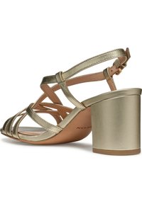 Sandal de tacón alto dorado metálico con tiras entrecruzadas, punta abierta y un tacón grueso con una textura suave y una plantilla marrón suave.