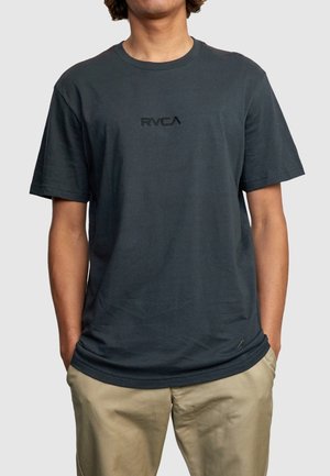 Personne portant un t-shirt noir à manches courtes avec le logo "RVCA" sur la poitrine et un pantalon beige, debout les mains dans les poches.