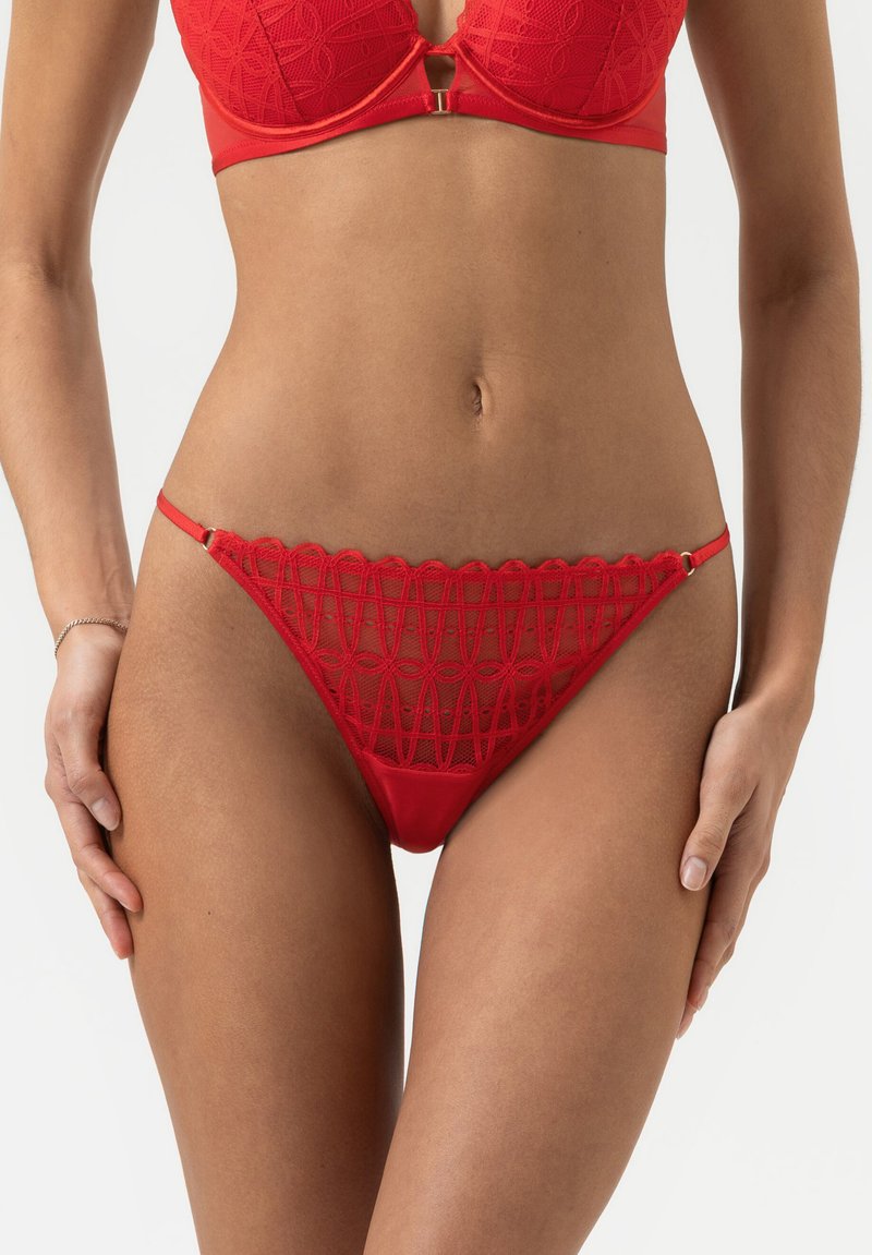 Rode kanten string met golvende randen, voorzien van een geometrisch patroon en dunne bandjes. Glad stof met minimale hardware-accenten.