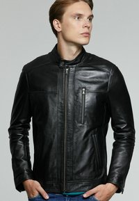 Chaqueta de cuero negra con cuello alzado, cuenta con bolsillos frontales con cremallera y costuras detalladas. Textura suave y diseño ajustado.