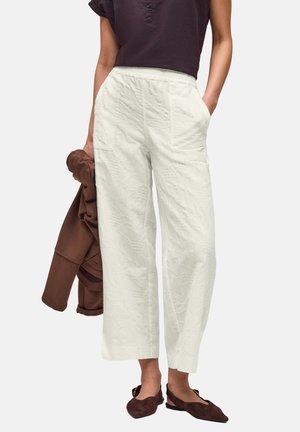 Kvinde iført hvide teksturerede bukser med høj talje og vide ben, mørkebrun top, holder en brun jakke og mørkebrun flad sko.