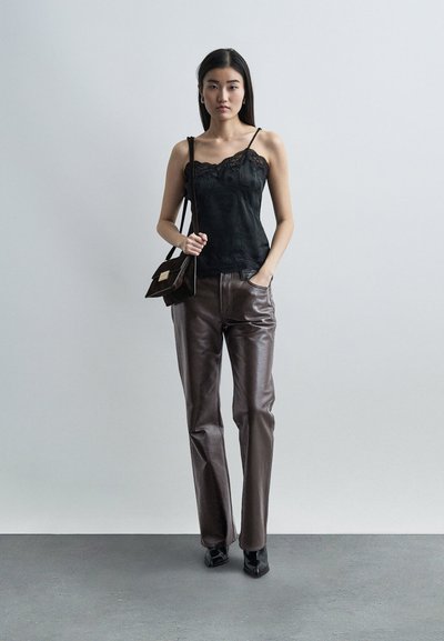 Mlada ženska stoji, oblečena v črno majico s čipkastim robom, rjave usnjene hlače, črne škornje in nosi rjavo torbico čez ramo.