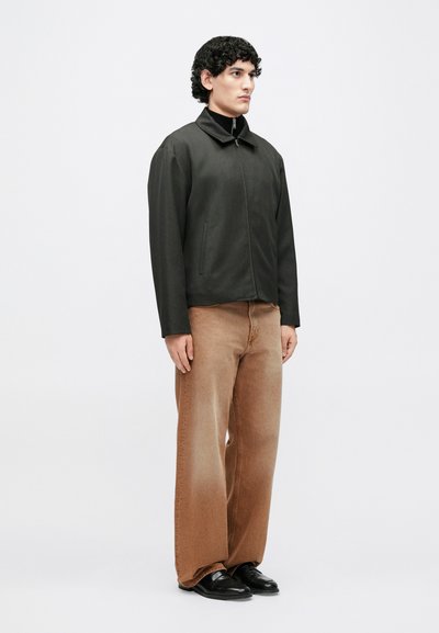 Zwarte jas met opstaande kraag, ritssluiting aan de voorkant en zijzakken; gecombineerd met lichtbruine wijde broekspijpen en zwarte schoenen. Soepele textuur.