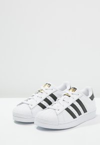 Sneakers bianchi con strisce nere, punta in gomma, design con lacci, dettagli dorati sulla linguetta e dettagli perforati ai lati.