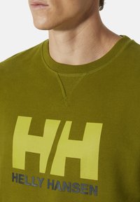 Zelená mikina s kulatým výstřihem vyrobená z bavlny, se světlým žlutým logem "HH" a textem "HELLY HANSEN" napsaným černě pod ním, s texturovanou tkaninou.