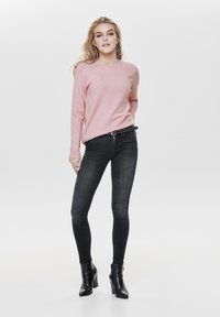 Rosa Strickpullover, schmale schwarze Jeans und schwarze Stiefeletten. Der Pullover hat einen runden Halsausschnitt und gerippte Bündchen; die Jeans sind eng und dunkel.