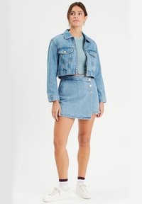 Veste en denim bleu coupée avec poches poitrine à boutons, assortie à une jupe portefeuille en denim bleu clair avec des boutons argentés et des baskets assorties.