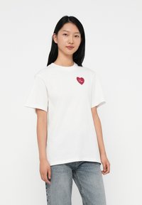 Camiseta de algodón blanca con mangas cortas, que presenta un corazón rojo bordado en el pecho con la palabra "Hugo" en una fuente decorativa.