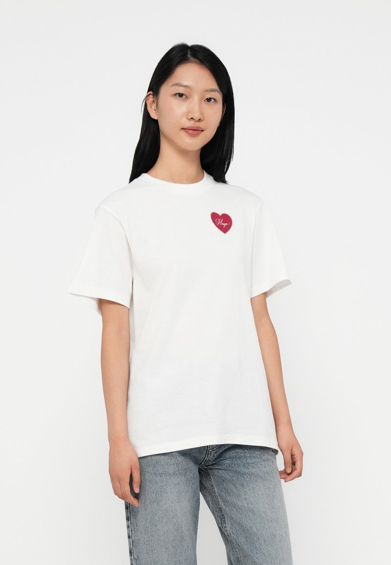 Camiseta de algodón blanca con mangas cortas, que presenta un corazón rojo bordado en el pecho con la palabra "Hugo" en una fuente decorativa.